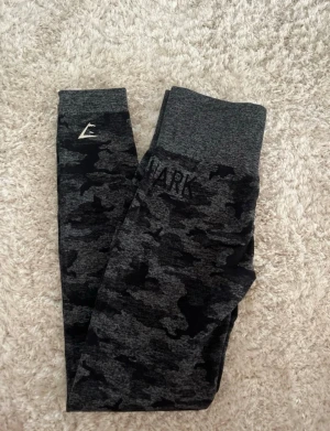 Svarta leggings i camo-mönster från Gymshark - Säljer ett par snygga tights från Gymshark i svart camo-mönster. De har hög midja och är tillverkade i ett stretchigt material som sitter skönt och följer kroppen. Perfekta för träning och gym. Gymshark-logga på benet + diskret text vid höften. Köpta i strl XS, passar även S då de töjer sig lite vid användning.