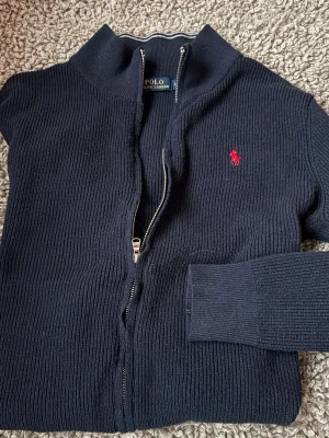 Marinblå stickad full zip Polo Ralph Lauren - Säljer en marinblå stickad kofta från Polo Ralph Lauren med dragkedja och ribbad struktur. Tröjan har lång ärm, hög krage med vita ränder och rött broderat logomärke på bröstet. Perfekt för dig som gillar klassisk stil med en sportig touch. Tröjan är storlek LARGE men sitter mer åt MEDIUM hållet.