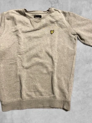 Grå sweatshirt från Lyle & Scott - En stilren grå sweatshirt från Lyle & Scott Junior med klassisk rund hals och det ikoniska gula logomärket broderat på bröstet. Tröjan har långa ärmar och ribbade muddar vid ärmslut och nederkant. Perfekt för en avslappnad look. Storlek 10-11 years. Har en liten svart fläck på magen som knappt syns!