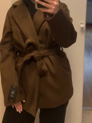 Brun kappa med bälte från NA-KD - Säljer en brun kappa från NA-KD i storlek 34. Jackan har en klassisk krage, knytbälte i midjan och är långärmad. Stor i storlek och passar även m beroenden på hur man vill att den ska sitta!! 