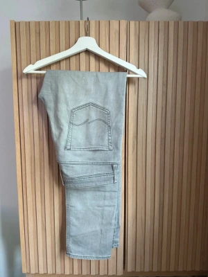 Grå jeans från Jack & Jones, 33/34 - Säljer ett par ljusgrå jeans från Jack & Jones i storlek 33/34. Klassisk femficksmodell med raka ben och snygga sömmar på bakfickan. Jeansen är tillverkade i mjukt bomullsmaterial och har en stilren look som funkar till många olika outfits.