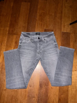 Grå jeans från Jack & Jones, 28/30 - Snygga grå jeans från Jack & Jones i klassisk femficksmodell. Jeansen har en rak passform och är tillverkade i mjukt bomullsmaterial med lite stretch. Perfekta för dig som gillar en stilren look med ljusgrå tvätt och diskreta detaljer.