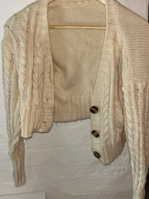 Beige croppad stickad kofta - Säljer en beige croppad stickad kofta med flätstickade detaljer och stora bruna knappar framtill. Koftan har lång ärm och bred ribbad mudd vid ärmslut och nederkant. Perfekt för lager på lager under höst och vinter.
