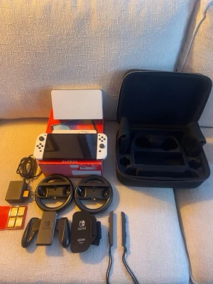 Nintendo Switch OLED Spelkonsol vit med tillbehör och spel - Nintendo Switch OLED spelkonsol i vit färg. Paketet inkluderar konsol med OLED-skärm, laddare, två rattar, Joy-Con-kontroller, förvaringsväska, samt fyra spel: Mario Kart 8 Deluxe, Super Mario Odyssey, Nintendo Switch Sports och 51 Worldwide Games. Dessutom medföljer ett SanDisk microSD-kort på 128 GB för extra lagringsutrymme.