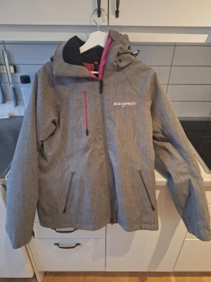 Grå vindjacka från Everest med huva stl 40 - Säljer en grå vindjacka från Everest med snygga rosa detaljer på dragkedjorna och insidan. Jackan har huva, tre fickor med dragkedja och en sportig look. Perfekt för dig som gillar att vara ute och vill ha en jacka som är både praktisk och stilren.