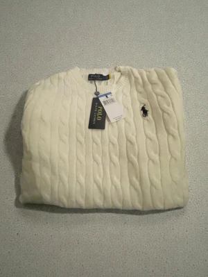 Vit kabelstickad tröja Polo Ralph Lauren - Snygg vit kabelstickad tröja från Polo Ralph Lauren i storlek S. Tröjan har rund hals, långärmade ribbade muddar och klassisk broderad logga på bröstet. Tillverkad i 100% bomull för en mjuk och skön känsla. Perfekt för dig som gillar stilrena och tidlösa plagg. Visst, här är några hashtags och sökord för bilden:  Hashtags: #RalphLauren #Polo #Sweater #Cream #CableKnit #Fashion #Style #Clothing #New #WinterFashion  Ralph Lauren, Polo, tröja, cream, kabelstickad, ny, kläder, mode, stil.
