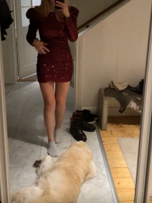 Vinröd glittrig kortklänning - Säljer en vinröd kortklänning med långa ärmar och axelvaddar. Klänningen är täckt av glittriga paljetter och har en öppen rygg som ger en snygg detalj. Materialet känns festligt och figurnära. Den är i nytt skick. Hör av er vid funderingar💗