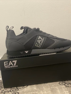 Emporio Armani EA7 svarta sneakers - Säljer ett par stilrena svarta EA7 Emporio Armani sneakers med meshpaneler och snygga detaljer i silver. Skorna har snörning, platt sula med räfflad undersida och logga på sidan. Perfekta för dig som gillar sportig och modern design. Priset går att diskutera.
