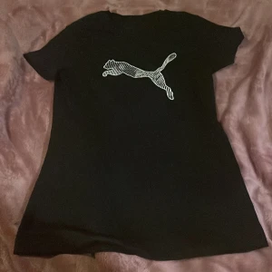 Svart Puma t-shirt med logga - Svart t-shirt från Puma med stor vit logga i form av en hoppande puma på bröstet. Klassisk rund hals och korta ärmar. Materialet är mjuk bomull som känns skönt mot huden. Perfekt för dig som gillar sportiga och stilrena plagg.Endast använd fåtal gånger.