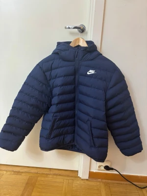 Mörkblå pufferjacka från Nike XL - Säljer en mörkblå pufferjacka från Nike i storlek XL (158-170 cm) helt ny. Jackan har huva, dragkedja framtill och två fickor på sidorna. Klassisk Nike-logga på bröstet och quiltad design som håller värmen. Perfekt för kalla dagar.
