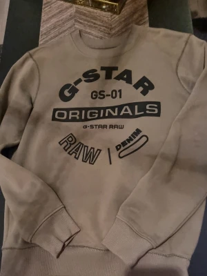 Grön sweatshirt från G-Star Raw M - Säljer en grön sweatshirt från G-Star Raw i storlek M. Tröjan har svart tryck framtill med texten 'G-STAR ORIGINALS RAW DENIM'. Klassisk rund halsringning och ribbade muddar vid ärmslut och nederkant. Perfekt för en chill och stilren look.