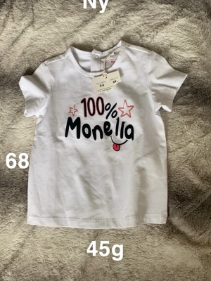 Vit t-shirt med tryck - Säljer en vit t-shirt med färgglatt tryck '100% Monella' och två röda stjärnor framtill. T-shirten har rund hals och korta ärmar. Perfekt för dig som gillar lekfull stil och bekväma plagg.🌸 Ny med lapp. Storlek 68