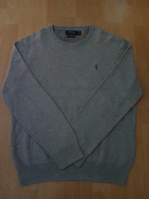 Grå stickad tröja från Polo Ralph Lauren. - Klassisk grå stickad tröja från Polo Ralph Lauren med rund halsringning och diskret broderad logga på bröstet. Tröjan har ribbade muddar vid ärmslut och nederkant, perfekt för lager på lager under höst och vinter. Bredd: 46cm / Längd: 61cm. Nypris: 2295 / Mitt pris: 799kr