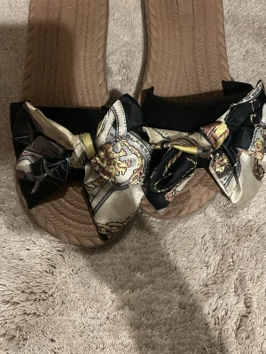Sandaler med mönstrad rosett - Säljer ett par snygga sandaler med beige sula och breda band i svart tyg dekorerade med stora, mönstrade rosetter i sidenliknande material. Perfekta för sommardagar och ger en trendig touch till din outfit. Storlek 39.