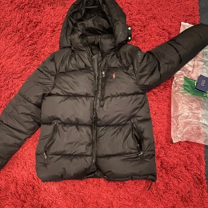 Svart pufferjacka från Polo Ralph Lauren - Säljer en svart pufferjacka från Polo Ralph Lauren med huva och dragkedjor framtill. Jackan har det klassiska röda Polo-logot broderat på bröstet och flera praktiska fickor med dragkedja. 