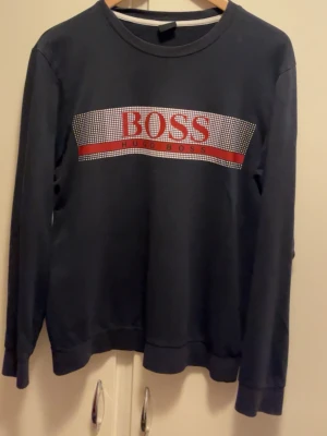Blå sweatshirt från Hugo Boss - Snygg blå sweatshirt från Hugo Boss med stort rött och vitt logotryck på bröstet. Tröjan har rund halsringning och långa ärmar, perfekt för en stilren och avslappnad look. Passar dig som gillar klassiska märken och enkel design.