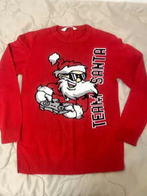 Röd jultröja med cool tomte - Röd stickad tröja med en stor bild av en tomte i solglasögon som håller i en spelkontroll och texten 'TEAM SANTA' på framsidan. Tröjan har rund hals och långa ärmar, perfekt för julens alla tillfällen.