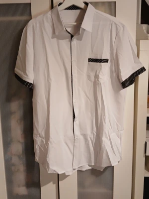 Vit skjorta med prickiga detaljer XL. Från Temu  - Snygg vit skjorta i storlek XL med svarta detaljer med vita prickar på ärmslut, insidan av kragen och fickan. Skjortan har korta ärmar, klassisk krage och knäppning framtill. Perfekt för dig som gillar stilrena plagg med en twist. Från Temu!