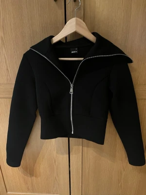 Svart croppad hoodie från Gina Tricot - Säljer en svart croppad hoodie från Gina Tricot med bred krage och silverfärgad dragkedja framtill. Tröjan har lång ärm och en clean, stilren look som passar perfekt till både jeans och kjol. Materialet är tjockt och mjukt för extra komfort.