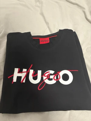Svart HUGO t-shirt med logga - Svart t-shirt från HUGO med stor vit och röd logga framtill. Klassisk rund halsringning och korta ärmar. Tillverkad i mjuk bomull som känns skön mot huden. Perfekt för dig som gillar stilrena plagg med tydlig branding.