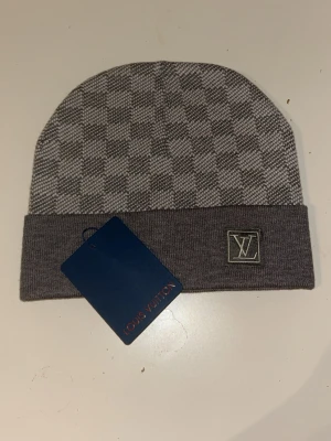 Grå rutig Louis Vuitton mössa - Säljer en snygg grå mössa från Louis Vuitton med klassiskt rutigt mönster och vikt kant. Mössan har en broderad LV-logga framtill och är stickad i mjukt material. Perfekt accessoar för dig som vill ha en exklusiv touch på din outfit. Finns kvitto