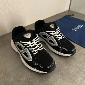 Svarta Dior b30. Mystery gift med på köpet! - Säljer ett par svarta Dior sneakers med stor vit CD-logga på sidan och vita snören. Skorna har en rund tå, platt sula och detaljer i syntet och skinn. Snygg kontrast mellan svart och vitt, perfekt för dig som gillar exklusiva märken och streetstyle.