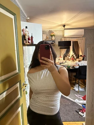 Vit prickig offshoulder topp - Säljer en vit topp med svarta prickar och offshoulder-design. Toppen är ärmlös och har en lätt rynkad effekt på sidan, vilket ger en snygg passform. Perfekt för dig som gillar en trendig och stilren look.