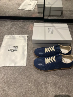 Blå Maison Margiela sneakers - Snygga blå sneakers från Maison Margiela med beige snörning och gummisula. Skorna har en mix av mocka och skinn på ovandelen och klassisk rund tå. Kommer med originalkartong och dustbag. Perfekta för dig som gillar stilrena och exklusiva sneakers.