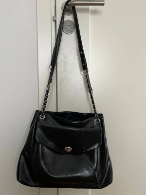 Svart axelväska med kedjedetaljer - Snygg svart axelväska i skinnimitation med långa axelband som har kedjedetaljer. Väskan har ett stort huvudfack och en framficka med vridlås. Perfekt storlek för att få plats med allt du behöver och ger en cool vibe till din outfit.