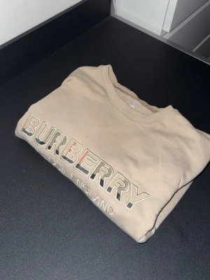 Beige sweatshirt från Burberry - Säljer en beige sweatshirt från Burberry med broderad logga i vitt, svart och rött på bröstet. Tröjan har rund halsringning, långa ärmar och ribbade muddar. Perfekt för dig som vill ha en clean och stilren look med exklusiv känsla.
