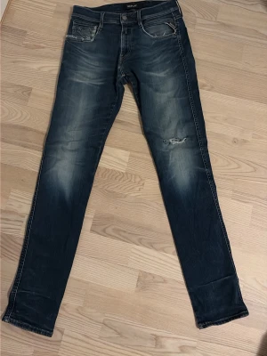 Replay anbass slim jeans - Säljer ett par blå skinny jeans från Replay med slitningar på ficka och knä. Jeansen har klassisk femficksdesign, snygga slitdetaljer och ljusare partier framtill. Materialet är jeans och passformen är tight med låg midja.