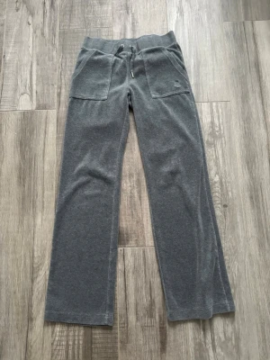 Mörkgrå mjukisbyxor med snörning - Säljer ett par mörkgrå mjukisbyxor från Juicy Couture med bred resår i midjan och snörning framtill. Byxorna har stora fickor fram och bak, samt broderad JC-logga på ena fickan. Sköna och perfekta för chill dagar.