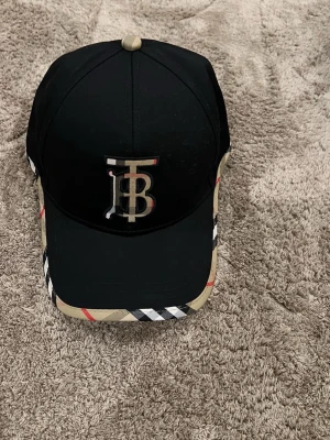 Svart keps med beige detaljer - Snygg svart keps med beige och rutiga detaljer samt ett stort TB-logotyp framtill. Perfekt accessoar för att lyfta din outfit och ge en sportig touch.