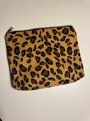 Leopardmönstrad sminkväska - Sminkväska med leopardmönster i beige, brunt och svart. Väskan har dragkedja i metall och är fodrad med vitt tyg på insidan. 