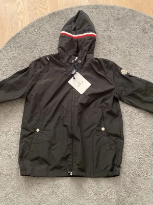 Svart vindjacka från Moncler med huva - Säljer en svart vindjacka från Moncler med huva och dragkedja. Jackan har vita och röda ränder på huvan, två fickor med tryckknappar och Moncler-logga på ärmen. 