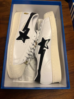 Adidas Superstar x BAPE sneakers - Säljer ett par Adidas Superstar x BAPE sneakers i vitt skinn med svarta detaljer och guldiga accenter. Skorna har klassiska tre svarta ränder på sidan, BAPE-stjärna och subtilt mönster i lädret. Tjock vit sula och snörning framtill. Riktigt clean och unik collab för dig som gillar streetwear.