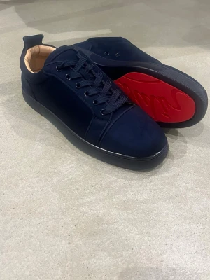 Mörkblå sneakers från Louboutin - Snygga mörkblå sneakers från Christian Louboutin med klassisk röd sula och diskret guldfärgad logga på insidan. Skorna har rund tå, snörning och ovandel i mocka. Insidan är beige och sulan är i gummi med signaturtext.