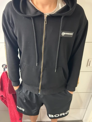 Svart Burberry hoodie med dragkedja - Svart hoodie från Burberry med dragkedja, huva och snören. Klassisk Burberry-ruta med logga på ena ärmen och Burberry-märke på bröstet. Mjuk bomullskänsla och känguruficka framtill. Snygg och stilren design med street-vibe. Ny pris: 3000+KR