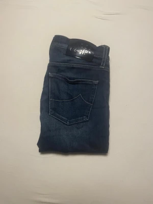 Jacob Cohën mörkblå jeans - Säljer ett par mörkblå jeans från Jacob Cohën med klassisk femficksdesign och snygga kontrastsömmar. Jeansen har en stilren läderpatch bak och silverfärgad knapp framtill. Materialet är mjukt jeans med exklusiv känsla och passformen är rak och normal.