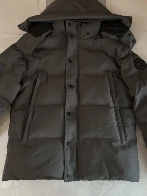 Grå pufferjacka från Canada Goose Label - Säljer en mörkgrå pufferjacka från Canada Goose label med huva och diskret logga på ärmen som man kan scana. Jackan har stora fickor, dragkedja och knappar framtill samt en riktigt schysst, vadderad look. Perfekt för kalla vinterdagar och riktigt snygg till streetwear.