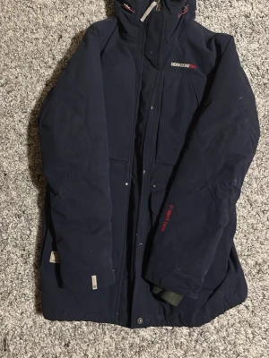 Mörkblå parkajacka Didriksons 1913 - Parkajacka från Didriksons 1913 i mörkblått med huva och dragkedja. Jackan har Storm System-foder i beige, praktiska fickor och diskreta röda detaljer. Perfekt för kalla dagar och har en lång, skyddande passform.