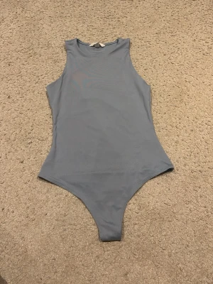 Ljusblå ärmlös bodysuit - Säljer en stilren ljusblå bodysuit med hög hals och ärmlös design. Toppen har knappar som man knäpper själv där nere