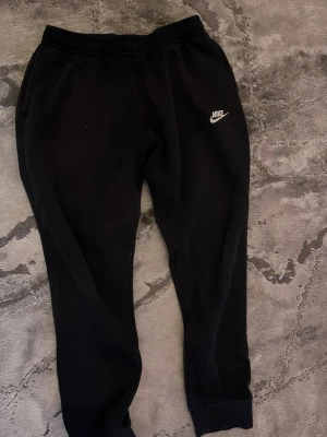 Svarta mjukisbyxor från Nike - Svarta mjukisbyxor från Nike med klassisk vit logga på vänster ben. Byxorna har resår i midjan och muddar vid bensluten för en skön passform. Perfekta för chill eller träning. Tillverkade i mjukt bomullsmaterial.