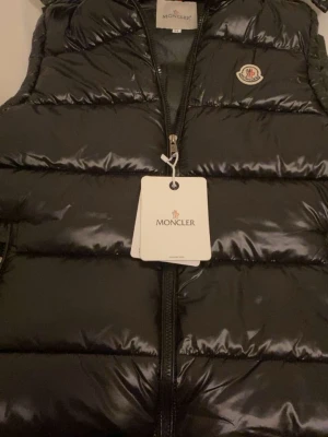 Svart dunväst från Moncler - Säljer en svart, glansig dunväst från Moncler med klassisk logga på bröstet. Västen har quiltad design, dragkedja framtill och coolt innerfoder med serietidningsmönster. Perfekt för lager-på-lager och riktigt snygg till streetwear-stilen.