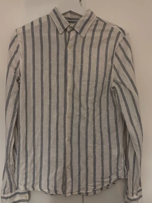 Randig linneskjorta från Dressmann  - Säljer en fräsch vit och blå randig skjorta från Dressmann i premium linne. Skjortan har klassisk krage, knäppning framtill och långa ärmar. Perfekt för dig som gillar en clean och avslappnad stil. Con 9/10. Använd endast fåtal gånger