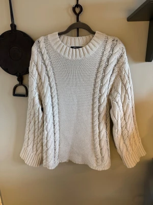 Vit stickad tröja från Gina Tricot - Säljer en chunky vit stickad tröja från Gina Tricot i storlek S. Tröjan har grova flätstickade detaljer längs ärmar och sidor, bred ribbad halsringning och muddar. Materialet är en mix av bomull och akryl, perfekt för kyliga dagar.