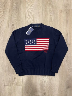 Mörkblå stickad tröja Polo Ralph Lauren - Mörkblå stickad tröja från Polo Ralph Lauren med amerikansk flagga i rött, vitt och blått på bröstet samt RL-broderi. Klassisk rund halsringning och ribbade muddar vid ärmar och nederkant. Perfekt för dig som gillar preppy stil.