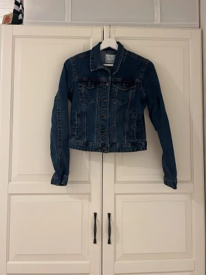 Mörkblå jeansjacka från Pull&Bear - Snygg mörkblå jeansjacka från Pull&Bear i klassisk modell med knappar framtill och två bröstfickor. Jackan har långa ärmar och coola kontrastsömmar som ger extra edge. Perfekt att slänga över en hoodie eller t-shirt för en chill look.