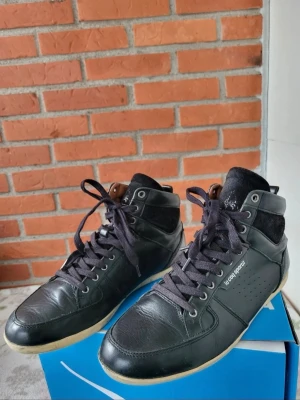 Svarta high top sneakers Le Coq Sportif 42 - Snygga svarta high top sneakers från Le Coq Sportif i skinn med svarta snören och detaljer i mocka. Skorna har perforerade sidor och en kontrasterande vit sula. Perfekta för dig som gillar en sportig och stilren look.