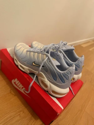Nike Air Max Plus vita/blå sneakers - Nike Air Max Plus sneakers i en fräsch kombination av vitt och ljusblått. Skorna har den ikoniska Tn Air-dämpningen, vågiga detaljer på ovandelen och en vit sula med synliga luftbubblor. Klassisk Nike Swoosh på sidan och Tn-logga bak. Perfekta för dig som gillar streetwear.